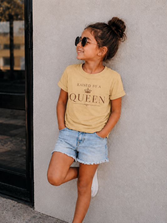 A young girl with her hair in a bun, wearing round sunglasses, a beige graphic t-shirt that reads 'RAISED BY A QUEEN', and denim shorts, posing against a textured gray wall.