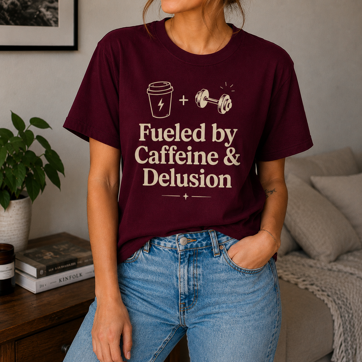 A woman wearing a maroon crewneck t-shirt with a cream graphic that reads 'Fueled by Caffeine & Delusion' with a coffee cup and dumbbell illustration, paired with blue jeans in a cozy living room setting.