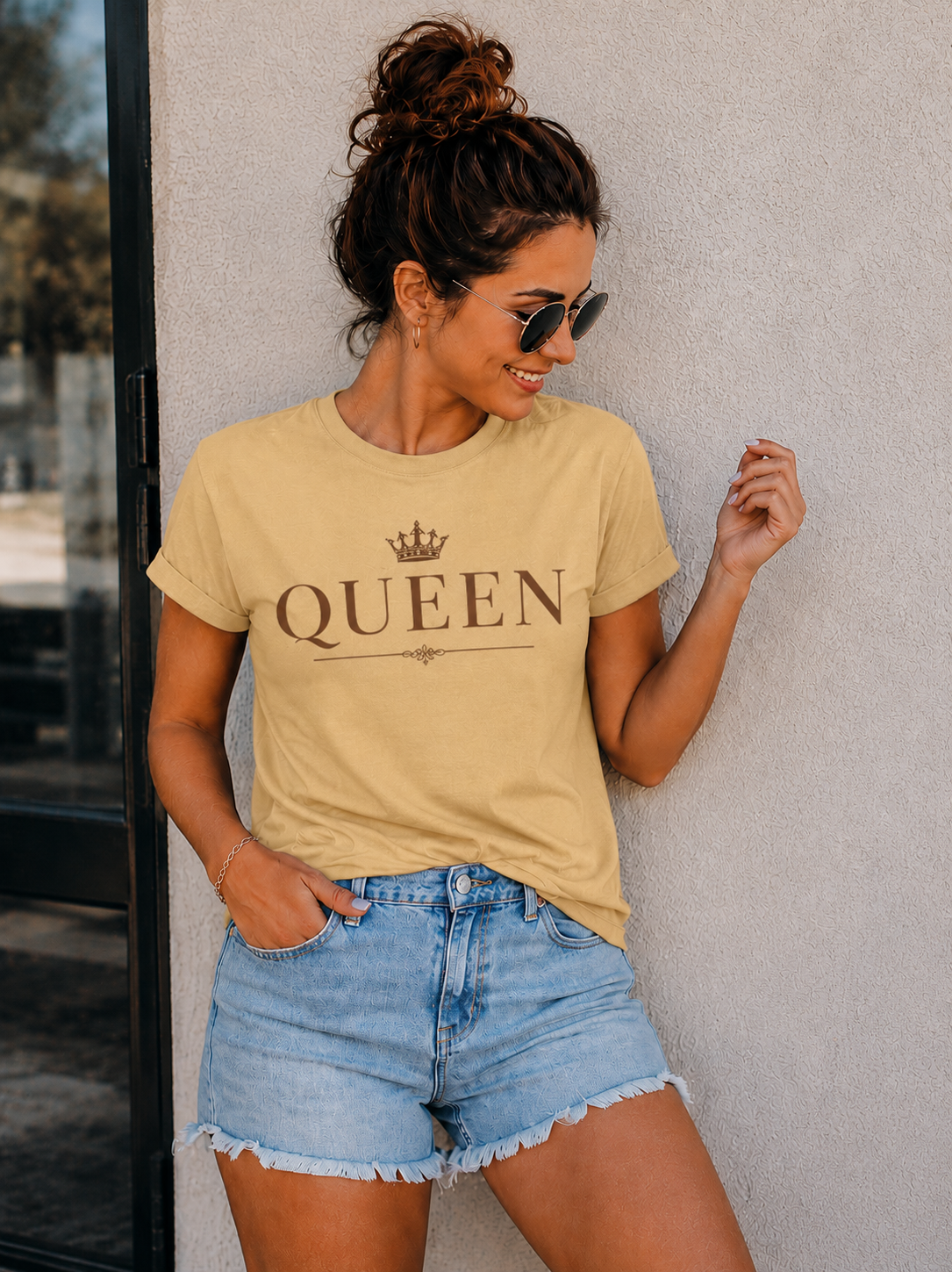 A woman wearing a beige t-shirt with a brown 'QUEEN' crown graphic and light blue denim shorts, posing against a textured gray wall.