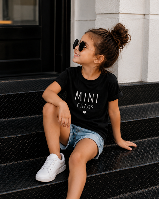 Young girl wearing the Mini Chaos black cotton graphic t-shirt with white typography, paired with denim shorts and white sneakers.