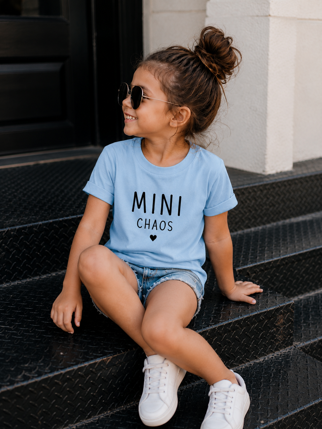 Young girl wearing the Mini Chaos blue cotton graphic t-shirt with white typography, paired with denim shorts and white sneakers.