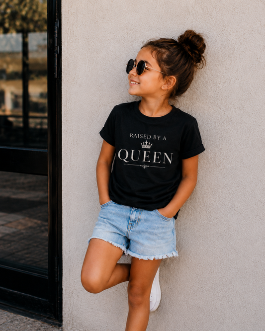 A young girl with her hair in a bun, wearing round sunglasses, a black graphic t-shirt that reads 'RAISED BY A QUEEN', and light blue denim shorts, posing against a textured gray wall.